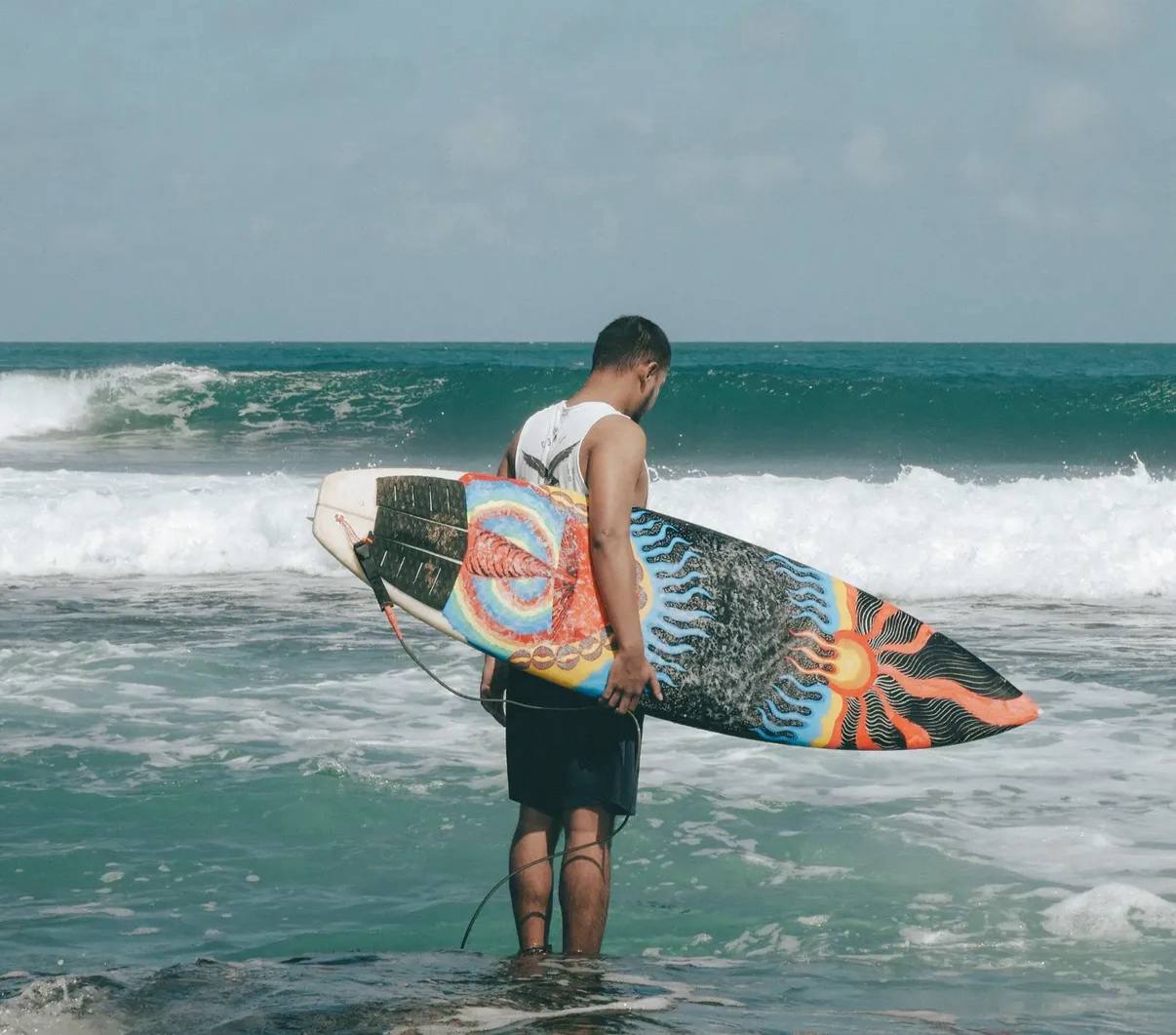 Man standing in shallow water holding a surfboard, waves behind