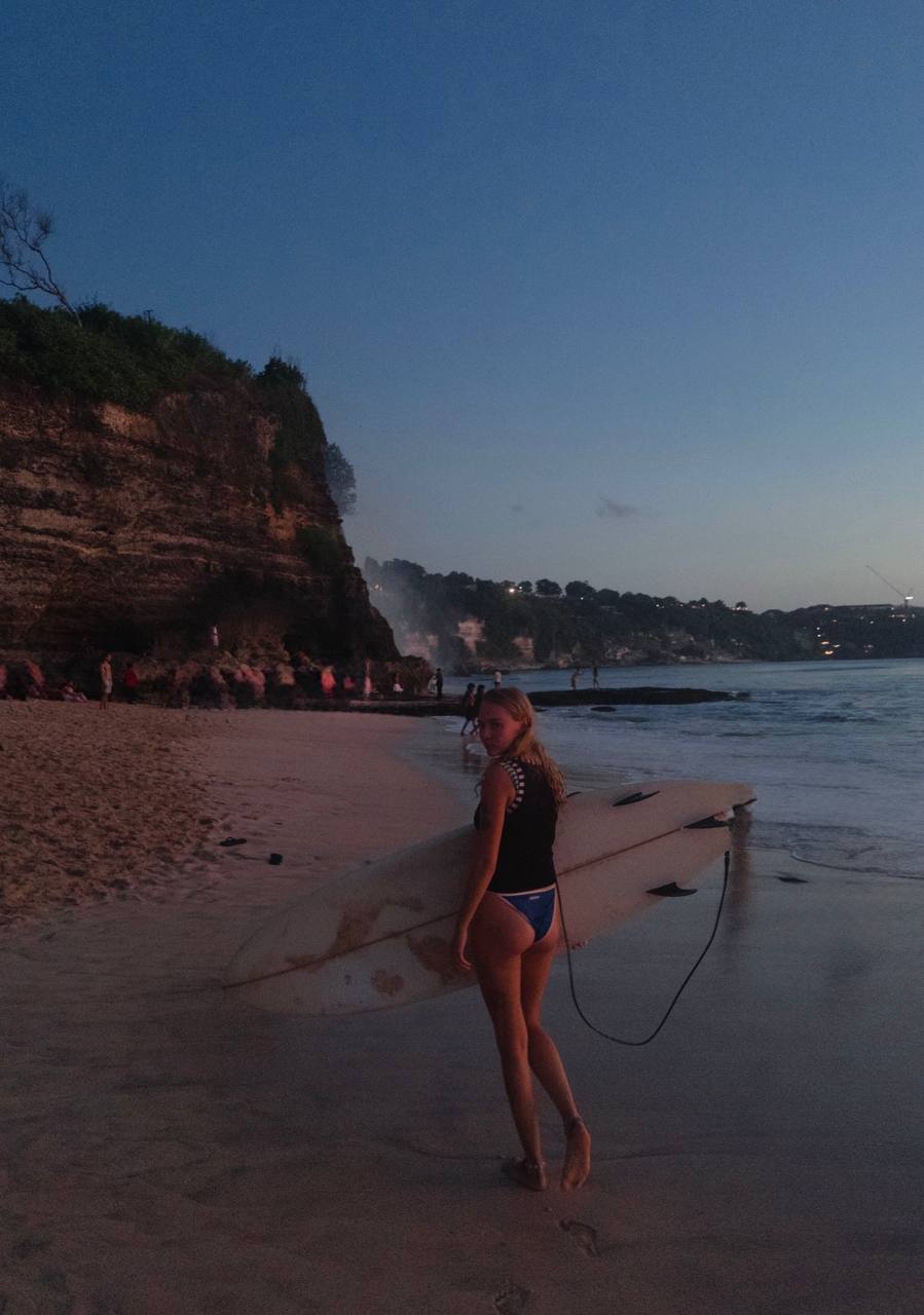 A woman carrying a surfboard on the beach at dusk, cliffs in the background